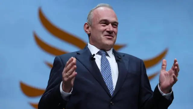 Liberal Democrat leader Ed Davey in a dark suit and blue tie. His hands are raised and he is smiling.