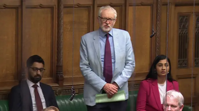 Jeremy Corbyn, a white man with short white hair, in a suit with red tie stood up in the House of Commons. He is standing in front of a green bench with other MPs sat next to him, a brown wall is behind him.