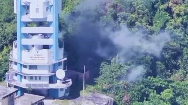 A tall hotel building next to some trees, you can see smoke rising from next to the structure and the trees