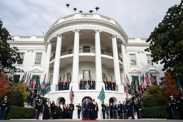 Trump and MBS standing on the red carpet in front of officials and US flags being held outside the White House