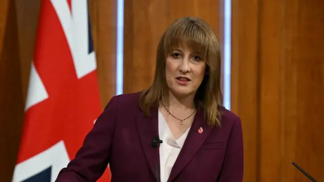 Chancellor Rachel Reeves wears a maroon jacket over a white blouse as she speaks at a media conference with the British flag draped behind her.