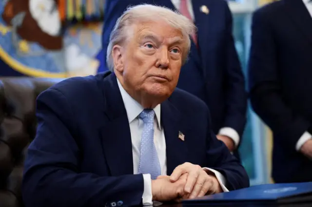 President Donald Trump sits at his desk in the Oval Office looking upwards with hands clasped while wearing a dark blue suite, light blue tie and white shirt