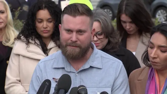 A bearded Sky Roberts wears a grey button-down shirt while he speaks to a media conference, with supporters behind him.