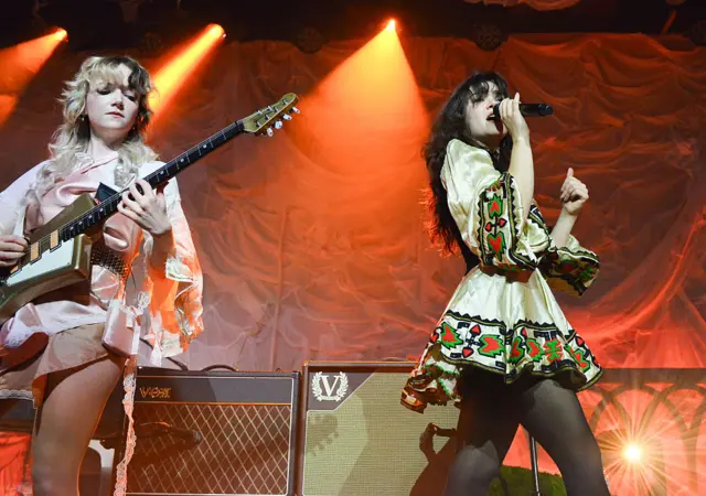A blonde woman playing a guitar and a woman singing into a microphone
