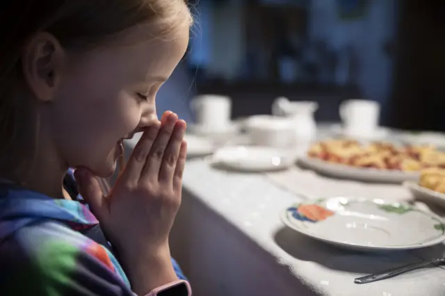 Young girl with blonde hair closes her eyes and puts her hands together in gesture of prayer at a dining table.