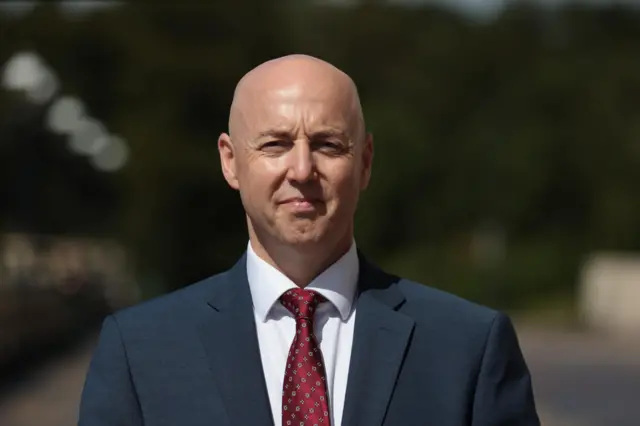A bald man in a navy suit, red tie, pictured outside