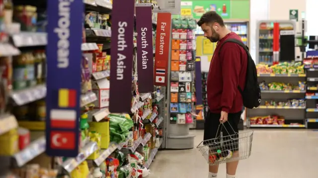 A man stands in a supermarket holding a basket and looks at the shelves