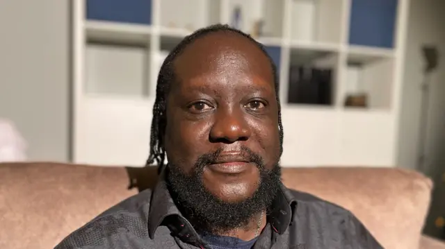 Henry Sabati McRae sitting on a pink sofa in his living room. Henry has brown eyes, longish hair and a beard. He wears a blue tee shirt and a black, textured, shirt