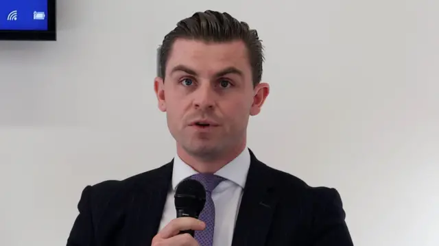 Darragh Mackin speaks into a handheld micrphone. He has slicked back, dark hair and is wearing a dark suit jacket, a white shirt and a purple tie.