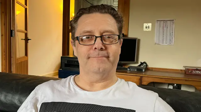 Mike Williams is sitting down on a sofa with a computer monitor behind him