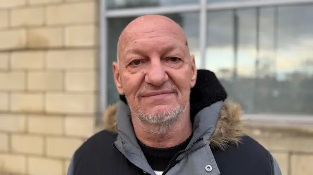 Michael has a is smiling, stood in front of the court in a black and grey jacket