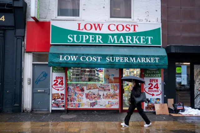 Person carrying umbrella walks past 'Low Cost Supermarket' on high street in rain