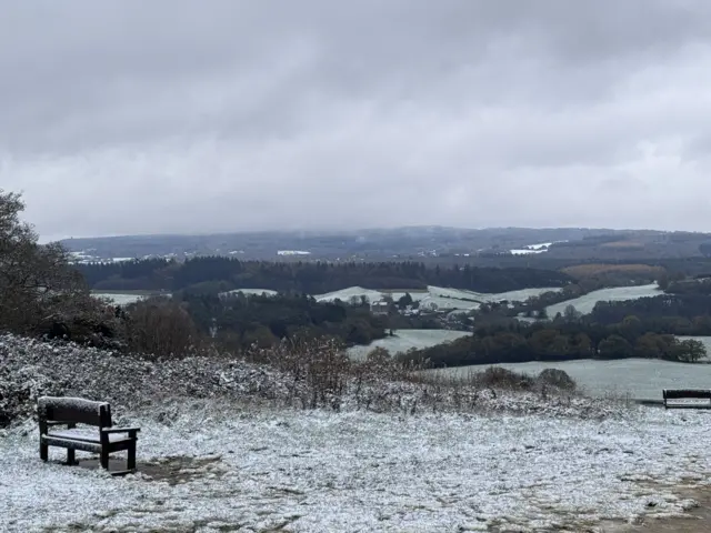 The top of a hill, with the ground and a bench covered in a light layer of snow, with the view beyond having a very light dusting