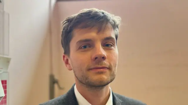 Billy Harding smiles at the camera inside a small room. He has short dark hair, some of which hangs over his forehead. He wears a dark grey suit with a white shirt and a tie.