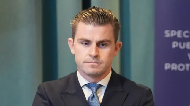 A close-up image of Darragh Mackin. He has slicked back, brown hair and he is wearing a dark suit jacket, a white shirt and a blue tie.