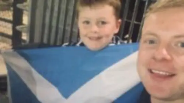 A man and a boy smiling at the camera. The boy is holding up a large Saltire flag
