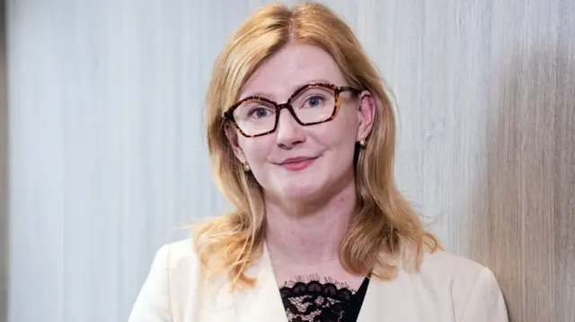 Karina Hutchens is standing in a corridor. She is wearing a cream blazer and a dark shirt. She has strawberry blonde hair and wears glasses