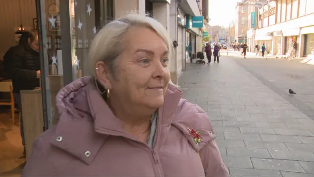 Donna has white blondish hair and stands in the street with a pink jacket on.