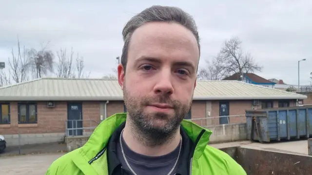 Anthony Flynn looks directly into the camera with a neutral expression. He has long, grey hair which is slicked back. He is standing in a yard with big containers and is wearing a green coat, a silver chain and a grey t-shirt.