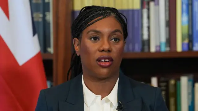 Kemi Badenoch wears a suit and white shirt sat in front of a union jack flag and a bookcase