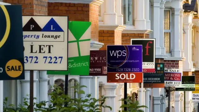 A row of houses with to let, for sale, and sold house signs on stands. The signs are from various different letting agencies and are a vast range of different colours. The houses are made from red brick and have white windows and doors.