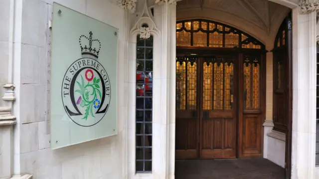 The exterior of the Supreme Court in London. There are large, wooden doors with stained glass windows. There is a sign displaying 'The Supreme Court' outside the grey stone building.