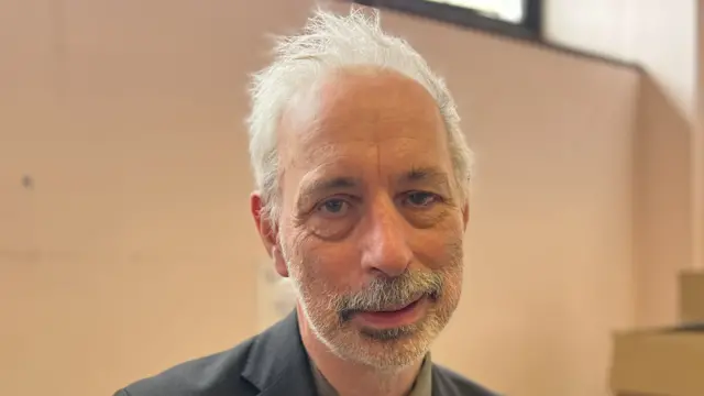Angus King smiles at the camera inside a room. He has short grey hair and a grey beard. He wears a navy blue suit with a dark green shirt underneath.