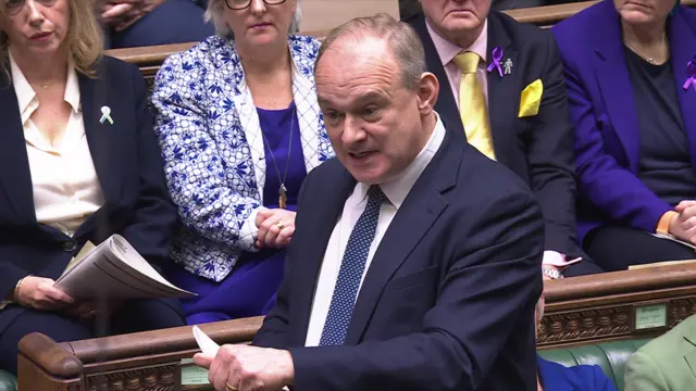 Ed Davey wearing a suit asks a question from a bench inside the House of Commons.