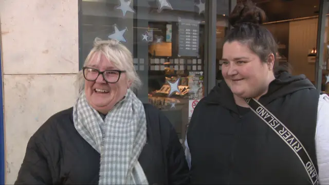 Cara has white blonde hair and is laughing. She wears glasses and a grey scarf. Fiona has her dark hair in a bun and is also smiling.