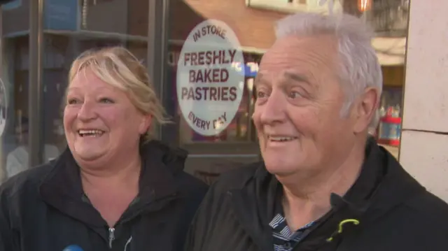 A blonde woman and a grey haired man stand smiling. Thye both look off camera while talking to a presenter. John holds a small brown furry dog in his hand.