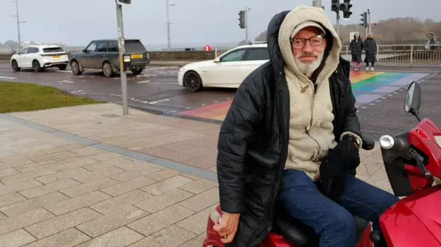 A man with glasses sits on a bench with a black coat and wooly cardigan under it