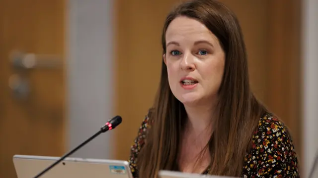 A woman with long brown hair is wearing a black open collar top speckled with red yellow and white. She is speaking into a thin mic, at a meeting of the Policing board. In front of her is an open silver laptop.