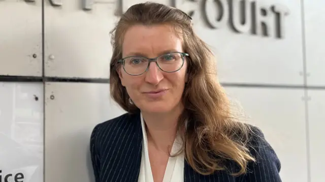 Niina Ankers, looks into camera with the county court sign behind her. She has long brown hair and is wearing glasses, a navy blazer and white blouse