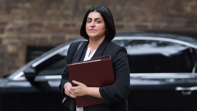 Shabana Mahmood in black blazer and white top walks towards No10 holding a burgundy folder, a car parked behind her