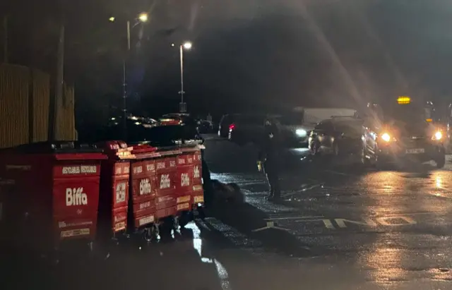A man is seen being arrested in a parking lot, There are bins beside him, as he lies face down on the ground and it appears to show authorities placing his hands in handcuffs