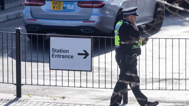 A single police officers walks past a sign reading Station Entrance while speaking on his shoulder-strapped radio