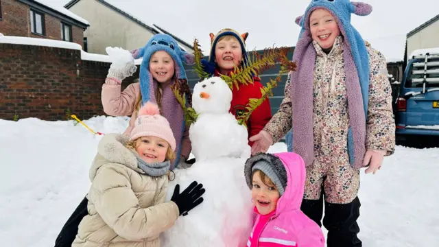 Five children surround a snowman that has been freshly built on a snowy drive.