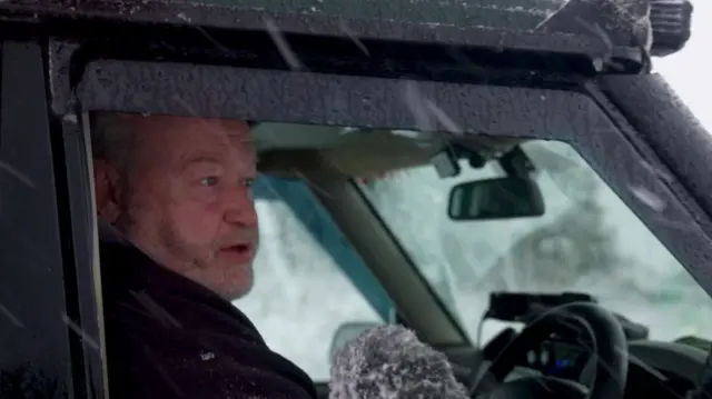 A man with a short grey stubble beard and moustache wearing a black fleece sat in a black car. A fluffy microphone is held close to him.