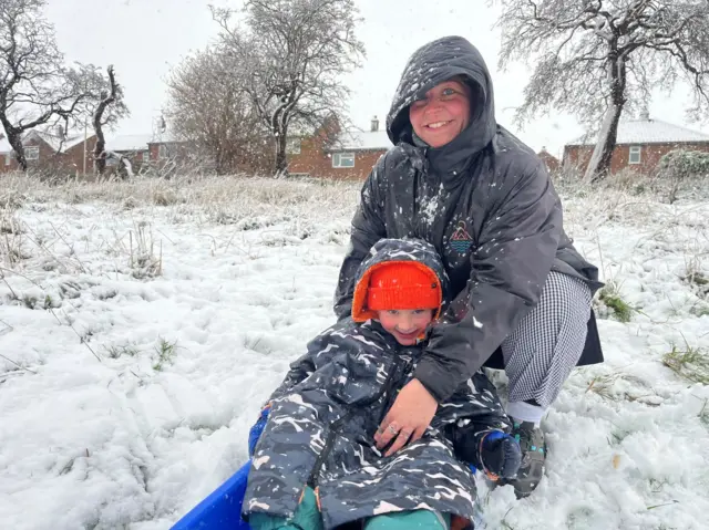 A woman and her son dressed in winter clothing pose in the snow. The boy is sitting in a blue sledge