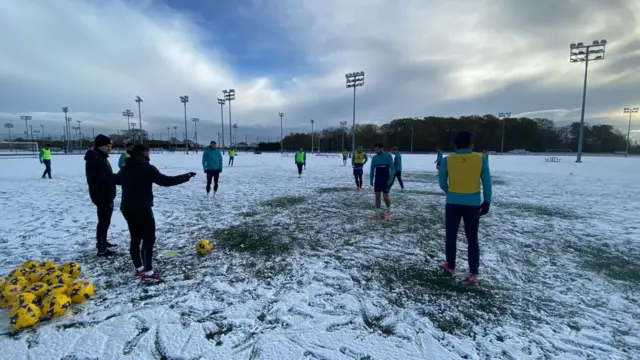Football players on a snow-covered pitch next to a large set of yellow balls
