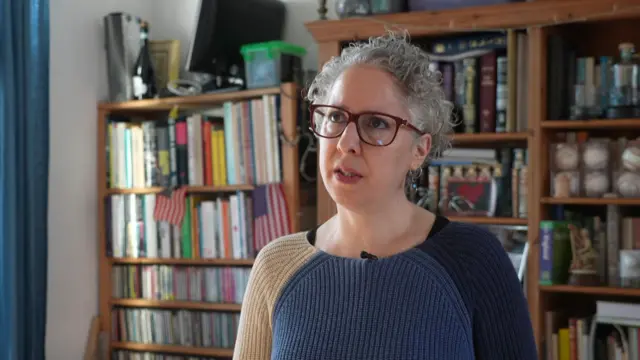 Rivka Gottlieb in her home. She is wearing a knitted jumper and has her hair tied back. There are bookshelves filled with books and photos and decorations in the background.