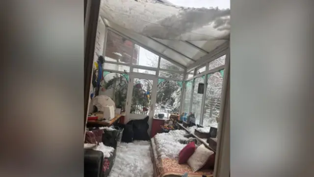 A conservatory attached to a house has suffered a roof collapse due to heavy snow. Transparent roof panels have given way, allowing snow to spill inside and cover the floor, furniture, and belongings.