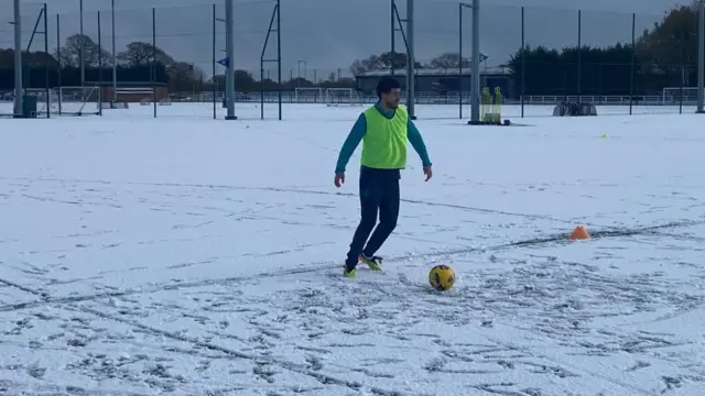 Footballer on a snow covered pitch playing with a yellow ball.