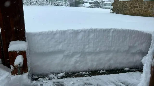 A thick layer of snow surrounds the entrance of a building. The snow has been neatly cleared from the doorway, revealing a vertical edge that highlights its depth—approximately a foot or more.