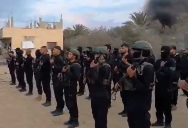A group of armed men are lined up in ranks holding rifles. Some have their faces covered. Most are wearing black combat uniforms