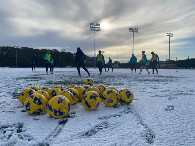 Several yellow footballs lay on a snowy pitch while players train in the background.