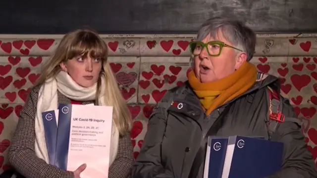 Brenda Doherty speaks in front of the Covid 19 memorial, which is covered in love hearts. She's carrying two physical copies of the inquiry's findings.