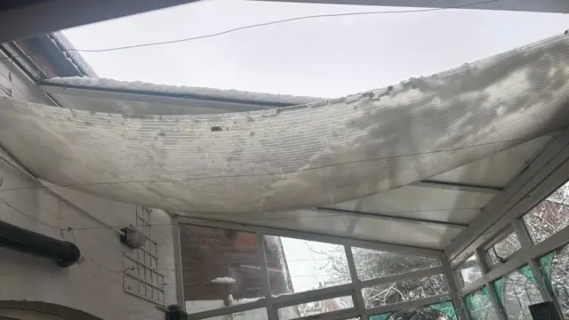 A conservatory with a translucent corrugated plastic roof shows signs of partial collapse due to heavy snow. The roof panels sag inward under the weight, forming a concave shape.