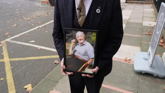 A man in a suit holds a photo of an elderly man, who is smiling to the camera.