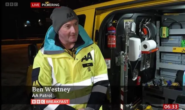 A man with a grey AA beanie and a yellow high vis AA coat looking to the side with BBC signage. It says he is live from Pickering and he is stood next to an open van.